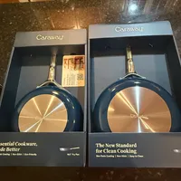 Customer-uploaded image. Click to see more details. The image represents: Two Caraway brand cookware boxes displayed on a dark granite countertop. The left box shows a 10.5-inch fry pan with blue exterior and gold interior, labeled "Essential Cookware, Made Better." The right box displays a similar fry pan with the tagline "The New Standard for Clean Cooking." Both boxes feature the Caraway branding and highlight non-toxic, non-stick, eco-friendly features.