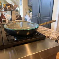 Customer-uploaded image. Click to see more details. The image represents: A black frying pan with glass lid sits on a stovetop in a modern kitchen with blue cabinets and granite countertops