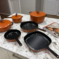 Customer-uploaded image. Click to see more details. The image represents: A set of orange and black cookware including pots with lids, frying pans, and a grill pan displayed on a marble kitchen counter with a cookbook