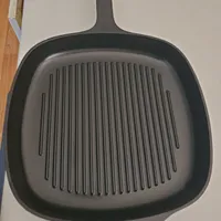 Customer-uploaded image. Click to see more details. The image represents: A square-shaped grill pan with raised ridges on the cooking surface and a single handle, photographed from above on a light-colored surface.