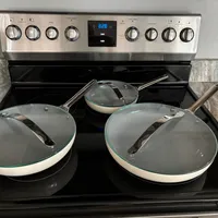 Customer-uploaded image. Click to see more details. The image represents: A stainless steel electric range with black cooktop surface displaying three cookware items: two large cream-colored skillets with glass lids on the front burners, and one smaller skillet with a glass lid in the back. The oven control panel shows a digital display reading 8:28, with multiple knobs for oven mode, temperature, and burner settings on either side.
