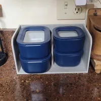 Customer-uploaded image. Click to see more details. The image represents: A white storage organizer on a granite countertop containing four blue stackable containers with white lids. The organizer is positioned against a beige wall with an electrical outlet and switch visible above it.