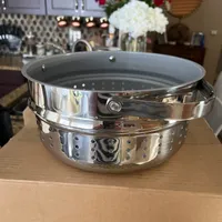 Customer-uploaded image. Click to see more details. The image represents: A stainless steel steamer basket or colander with perforated sides sitting on a kitchen counter, with kitchen cabinets and flowers visible in the background