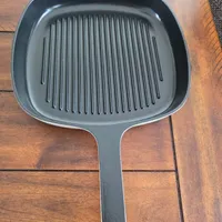 Customer-uploaded image. Click to see more details. The image represents: A black grill pan with raised ridges and a long handle, sitting on a wooden floor