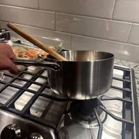 Customer-uploaded image. Click to see more details. The image represents: A stainless steel saucepan with a wooden handle sits on a gas stovetop in a kitchen. A wooden spoon rests in the pan. In the background, there's a subway tile backsplash and a skillet with food cooking on another burner.
