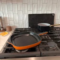 Customer-uploaded image. Click to see more details. The image represents: A black and orange grill pan sits on a gas stovetop in a modern kitchen with white subway tile backsplash and stainless steel countertops