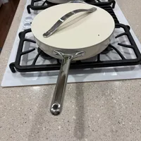 Customer-uploaded image. Click to see more details. The image represents: A cream-colored skillet with lid sits on a gas stovetop burner, with a metal spatula resting on the skillet. A large pot is visible in the background on another burner.