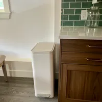 Customer-uploaded image. Click to see more details. The image represents: A modern kitchen with wood cabinets, green subway tile backsplash, white countertops, and a tall white step-on trash can positioned next to the counter