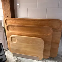 Customer-uploaded image. Click to see more details. The image represents: A collection of wooden cutting boards of varying sizes arranged in a leaning stack on a concrete floor against a white subway tile backsplash in a kitchen.