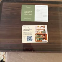 Customer-uploaded image. Click to see more details. The image represents: A wooden cutting board with Caraway brand informational cards including prep tips and warranty activation with QR code, placed on cardboard packaging