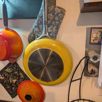 Customer-uploaded image. Click to see more details. The image represents: A kitchen wall display featuring colorful cookware including a yellow frying pan with stainless steel bottom, red-orange cast iron pots, a patterned kitchen towel, vintage kitchen signage, control dials, and a wire basket with lemons at the bottom.