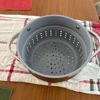 Customer-uploaded image. Click to see more details. The image represents: A gray colander with perforated holes and handles sitting on a striped kitchen towel on a wooden table