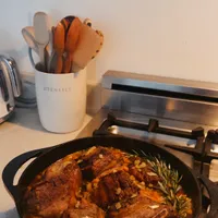 Customer-uploaded image. Click to see more details. The image represents: A cast iron skillet containing seared meat with herbs sits on a stovetop, with kitchen utensils in a container and a toaster oven visible in the background