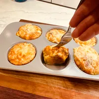 Customer-uploaded image. Click to see more details. The image represents: A muffin tin with six freshly baked golden-brown muffins, with someone's hand holding a fork near the center muffin. The tin sits on a wooden surface in front of a marble countertop with white subway tile backsplash and a blue glass container visible in the background.