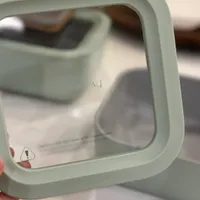 Customer-uploaded image. Click to see more details. The image represents: A pale green or sage-colored square mirror or reflective device being held up, showing a reflection of a white surface and blurred background elements.
