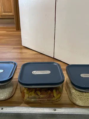 Customer-uploaded image. Click to see more details. The image represents: Three clear plastic storage containers with dark blue lids arranged on a wooden shelf. The left container holds white rice, the middle container holds pasta, and the right container holds oats or similar grain. Each lid has a label for identifying contents.