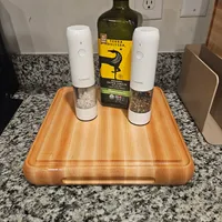 Customer-uploaded image. Click to see more details. The image represents: A wooden cutting board on a granite countertop with two white electric salt and pepper grinders and a bottle of Terra Delyssa olive oil arranged on top
