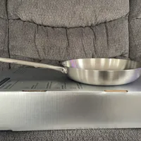 Customer-uploaded image. Click to see more details. The image represents: A stainless steel frying pan with long handle sitting on top of its white retail box on a gray couch