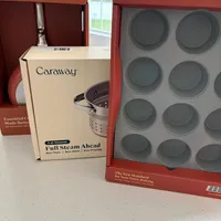 Customer-uploaded image. Click to see more details. The image represents: Caraway brand kitchen products displayed on a counter, including a boxed 3-quart steamer with non-toxic non-stick coating and a muffin tin with multiple circular cups, with red Caraway packaging boxes visible in the background near a window.