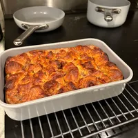 Customer-uploaded image. Click to see more details. The image represents: A freshly baked casserole with a golden-brown, bubbly top sits in a white rectangular baking dish on an oven rack. Two white ceramic bowls with metal handles and a metal pitcher are visible on the dark stovetop surface behind it.