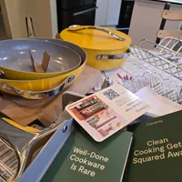 Customer-uploaded image. Click to see more details. The image represents: A kitchen counter displays yellow cookware including a skillet and covered pot, alongside green instruction booklets titled "Well-Done Cookware Is Rare" and "Clean Cooking Get Squared Away," with a magazine and wire basket visible in the background.