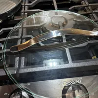 Customer-uploaded image. Click to see more details. The image represents: A stovetop with a glass lid resting on a dark pot, a metal tongs placed across the lid, a non-stick skillet visible on the left burner, and control knobs at the bottom showing heat settings from low to high.
