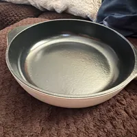 Customer-uploaded image. Click to see more details. The image represents: A round frying pan with a non-stick dark interior and light-colored exterior sitting on a brown textured surface with pillows in the background