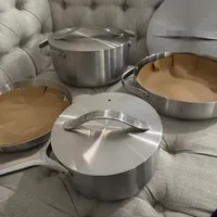 Customer-uploaded image. Click to see more details. The image represents: A set of stainless steel cookware including pots and pans with lids arranged on a light-colored tufted sofa or chair