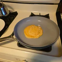 Customer-uploaded image. Click to see more details. The image represents: A golden pancake cooking in a gray non-stick skillet on a stovetop, with a kettle and plate visible in the background.