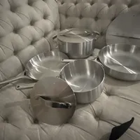 Customer-uploaded image. Click to see more details. The image represents: A set of stainless steel cookware including pots, pans and lids arranged on a tufted beige sofa or chair