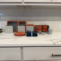 Customer-uploaded image. Click to see more details. The image represents: Kitchen counter with organized cookware including orange and gray dishes, bowls, cutting boards, and blue napkins displayed on white marble countertop with subway tile backsplash