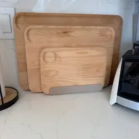 Customer-uploaded image. Click to see more details. The image represents: Three wooden cutting boards of graduated sizes arranged on a white marble countertop in a kitchen, with kitchen appliances and a white wall with light switch visible in the background.