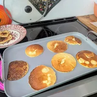 Customer-uploaded image. Click to see more details. The image represents: A griddle on a stovetop with eight pancakes in various stages of cooking, ranging from light golden to dark brown. A decorative pink plate with cooked pancakes sits nearby, along with colorful cookware and kitchen items in the background.