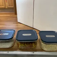 Customer-uploaded image. Click to see more details. The image represents: Three clear plastic storage containers with dark blue lids arranged on a wooden shelf. The left container holds white rice, the middle container holds pasta, and the right container holds oats or similar grain. Each lid has a label for identifying contents.