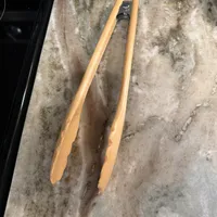 Customer-uploaded image. Click to see more details. The image represents: Wooden kitchen tongs with metal spring mechanism lying on a concrete floor next to a black container and white shoe.