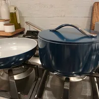 Customer-uploaded image. Click to see more details. The image represents: A kitchen stovetop with two dark blue enameled cast iron cookware pieces - a skillet on the left and a covered Dutch oven on the right, both sitting on burners. The background shows a white shiplap wall with wooden kitchen utensils and olive oil bottle on a wooden shelf.