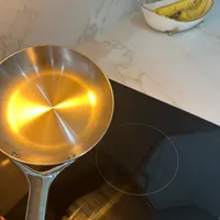 Customer-uploaded image. Click to see more details. The image represents: A stainless steel frying pan being held over an induction cooktop, with bananas visible in a bowl on the marble countertop in the background