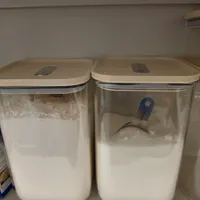 Customer-uploaded image. Click to see more details. The image represents: Two clear plastic storage containers with beige lids containing white powder or flour, positioned side by side on a shelf. The left container appears fuller than the right one, which has a blue measuring scoop inside.
