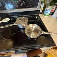 Customer-uploaded image. Click to see more details. The image represents: A stainless steel frying pan and saucepan sitting on a black glass-top electric stove, with cooking oils and ingredients visible on the counter nearby