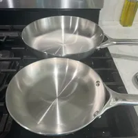 Customer-uploaded image. Click to see more details. The image represents: Two stainless steel frying pans sitting on a black gas stovetop in a kitchen