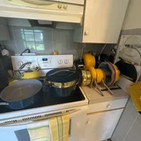 Customer-uploaded image. Click to see more details. The image represents: A compact kitchen with cream and gray cabinetry featuring a stovetop with various cookware including yellow and black pots and pans, a yellow kettle, a microwave above the stove, and gray tile backsplash with a window.