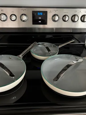 Customer-uploaded image. Click to see more details. The image represents: A stainless steel electric range with black cooktop surface displaying three cookware items: two large cream-colored skillets with glass lids on the front burners, and one smaller skillet with a glass lid in the back. The oven control panel shows a digital display reading 8:28, with multiple knobs for oven mode, temperature, and burner settings on either side.