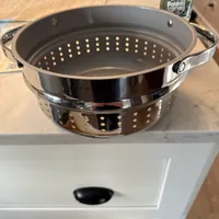 Customer-uploaded image. Click to see more details. The image represents: A stainless steel steamer basket with perforated holes sitting on a white kitchen counter, with handles on both sides and a Poland Spring water bottle visible in the background