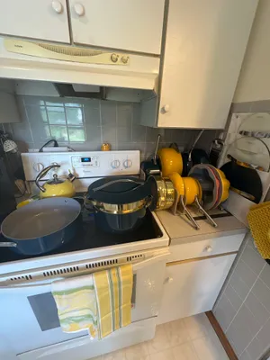 Customer-uploaded image. Click to see more details. The image represents: A compact kitchen with cream and gray cabinetry featuring a stovetop with various cookware including yellow and black pots and pans, a yellow kettle, a microwave above the stove, and gray tile backsplash with a window.