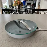Customer-uploaded image. Click to see more details. The image represents: A white ceramic frying pan with glass lid sits on a speckled granite kitchen counter, with a dining room and person in blue shirt visible in the background