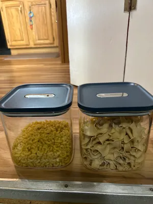 Customer-uploaded image. Click to see more details. The image represents: Two clear plastic storage containers with dark blue lids sit on a wooden shelf. The left container holds yellow cereal rings, and the right container holds tan-colored pasta shapes. Both containers have label slots on their lids. A wooden cabinet and white refrigerator are visible in the background.