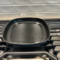 Customer-uploaded image. Click to see more details. The image represents: A black grill pan with ridged cooking surface sitting on a gas stovetop with stainless steel grates