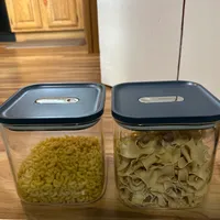Customer-uploaded image. Click to see more details. The image represents: Two clear plastic storage containers with dark blue lids sit on a wooden shelf. The left container holds yellow cereal rings, and the right container holds tan-colored pasta shapes. Both containers have label slots on their lids. A wooden cabinet and white refrigerator are visible in the background.