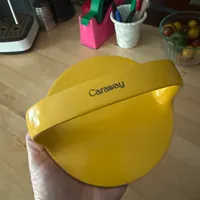 Customer-uploaded image. Click to see more details. The image represents: A hand holding a bright yellow Caraway brand mixing bowl with a pouring spout on a kitchen counter