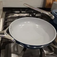 Customer-uploaded image. Click to see more details. The image represents: A white ceramic skillet with a dark blue exterior and stainless steel handle sits on a gas stovetop burner in a kitchen.